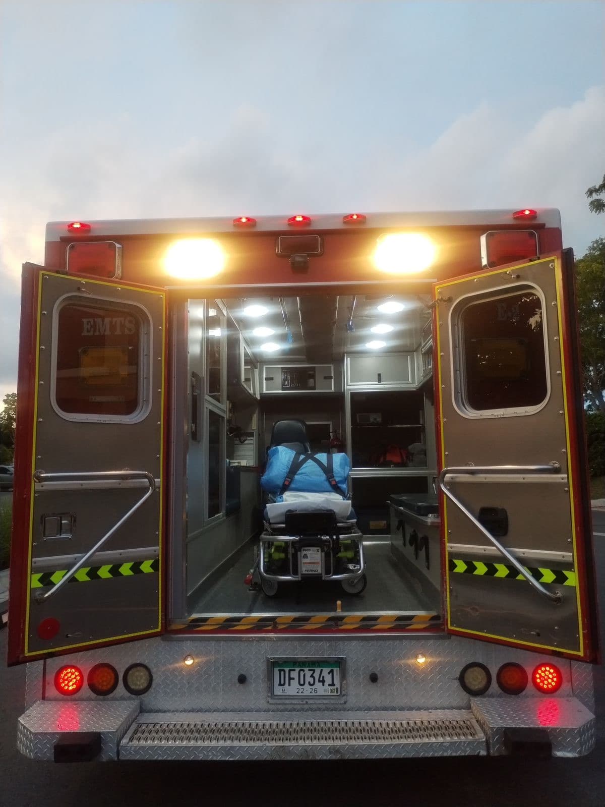An EMTS ambulance at dusk with its rear doors open, stretcher and equipment cabinets lit